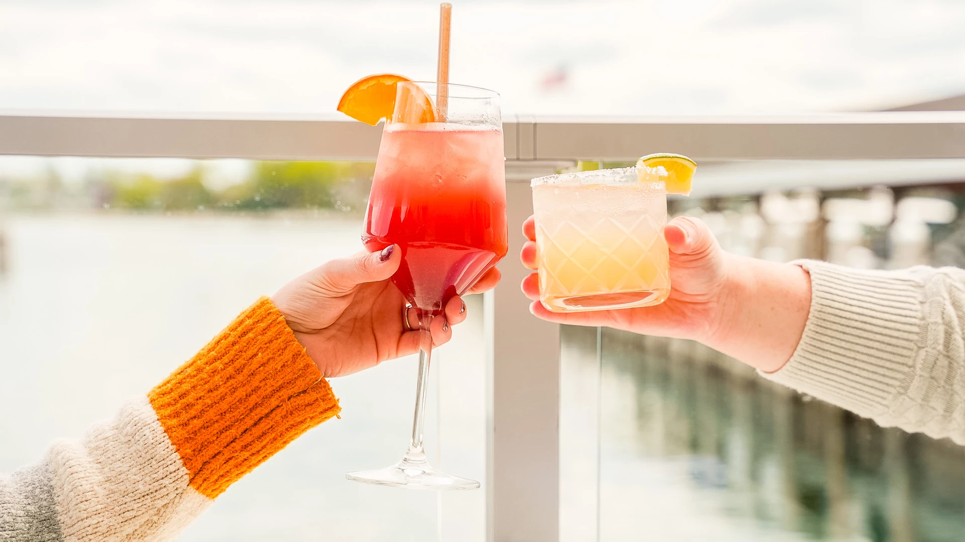 Two guests clinking a tall red cocktail and a salted citrus cocktail on the waterfront deck