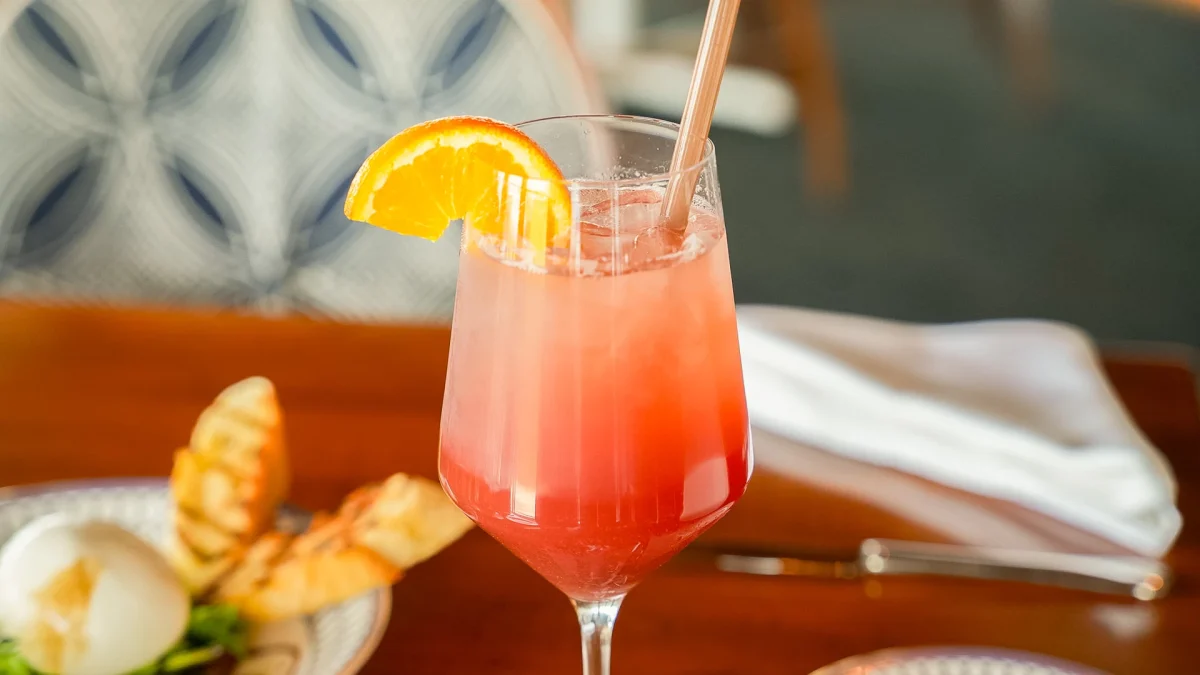 Pink sparkling cocktail with an orange slice and straw beside an appetizer plate
