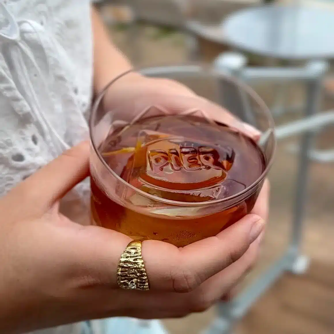 Hand holding an amber cocktail over a large ice cube stamped with the Pier logo and an orange twist