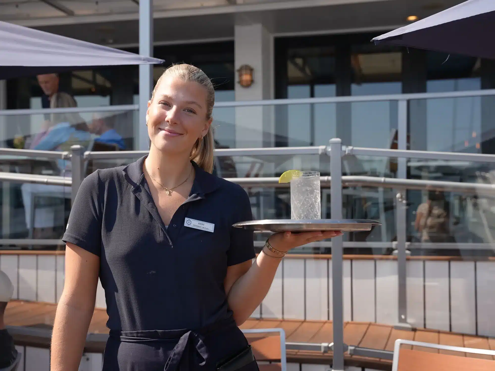 Smiling server holding a tray with a sparkling cocktail and lime wedge on the waterfront patio