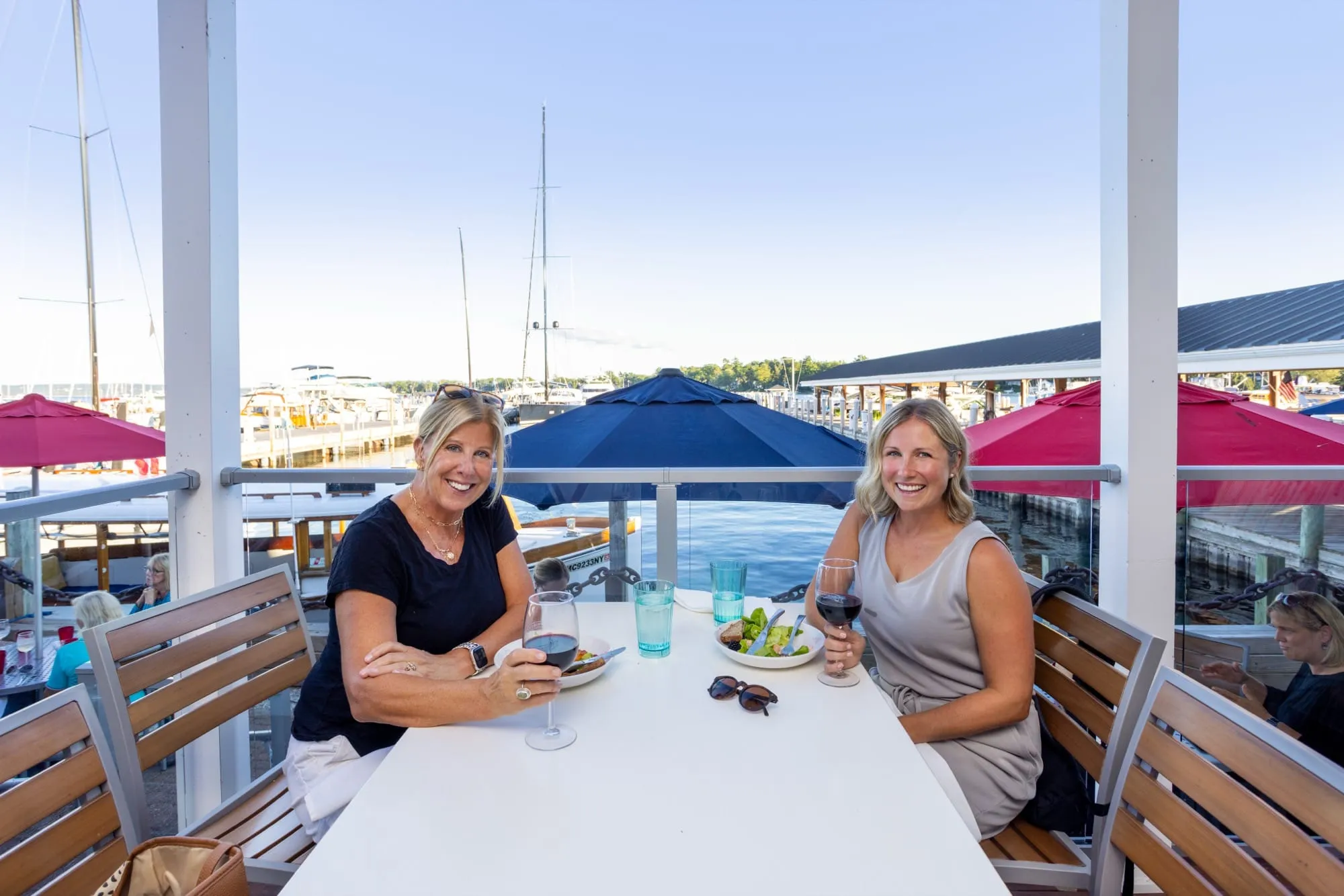 Two women smiling with glasses of red wine at a patio table overlooking the marina, with sailboats and red and navy umbrellas