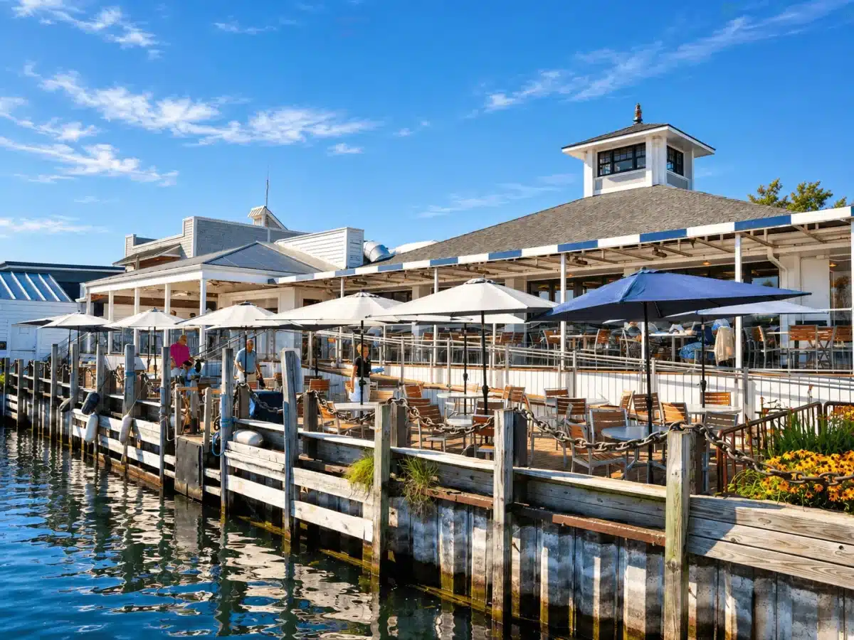 The restaurant's waterfront patio with white and navy umbrellas along the harbor dock, beneath the cupola-topped building on a sunny day
