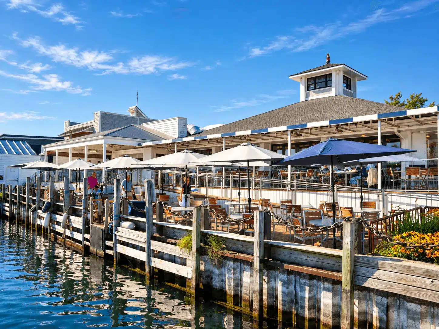The restaurant's waterfront patio with white and navy umbrellas along the harbor dock, beneath the cupola-topped building on a sunny day