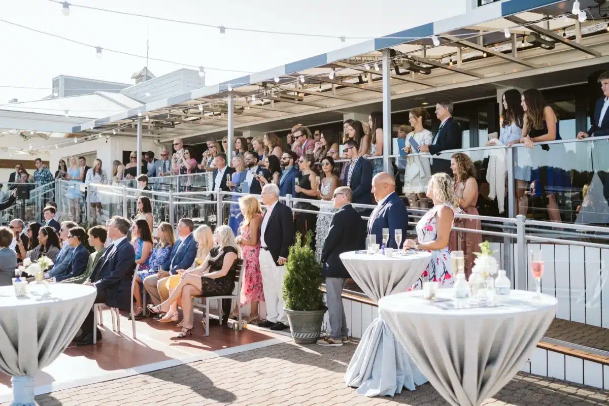 Wedding guests seated and standing on the deck and upper balcony, with high-top tables in gray linens and string lights overhead